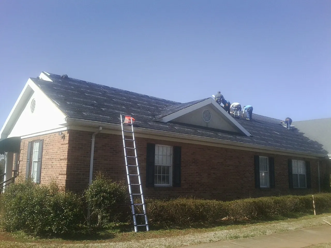Crew tearing shingles off a brick ranch roof with a ladder set — Emergency Roof Repair work by our crew in East Earl