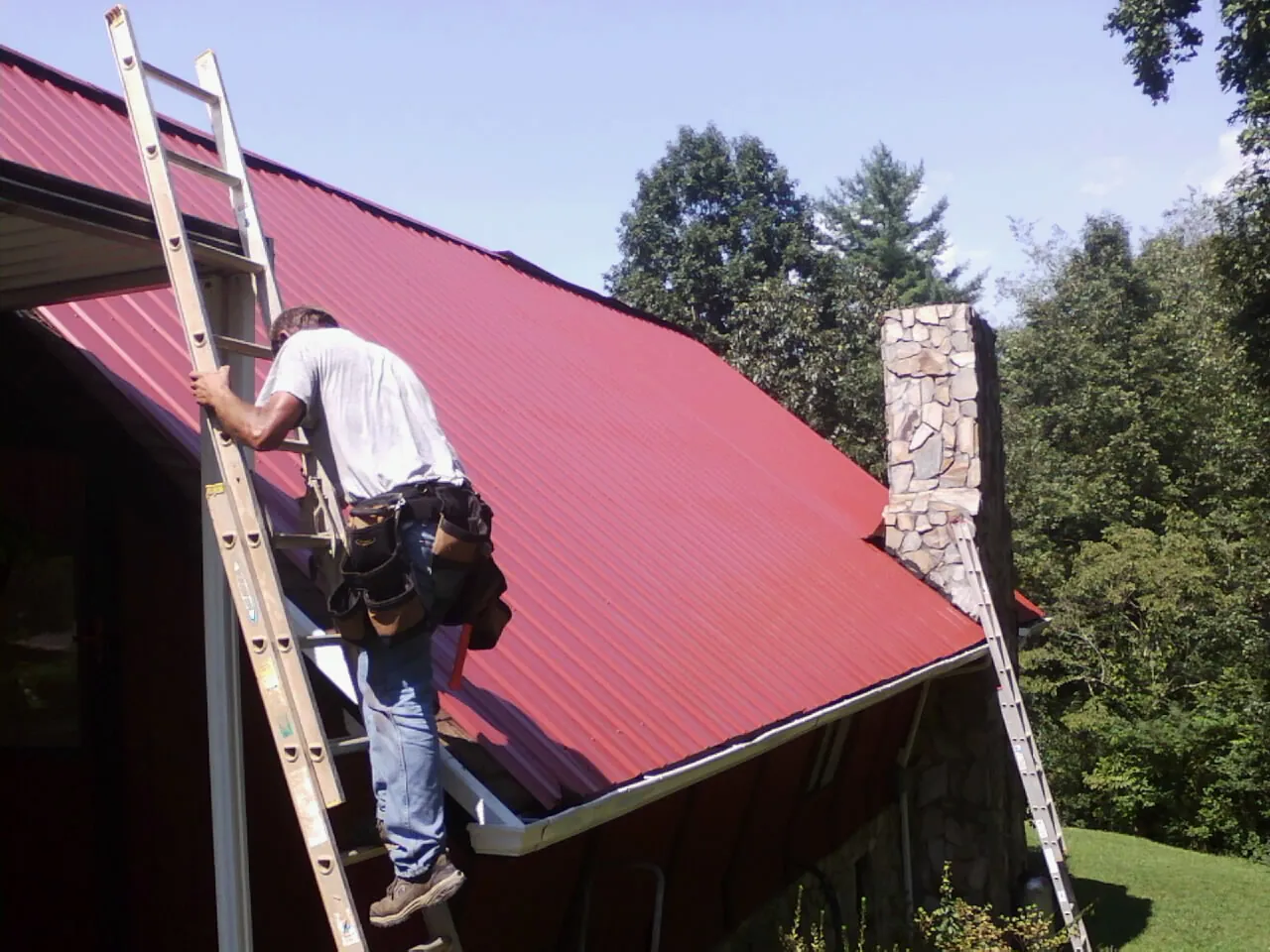 Roofer on a ladder beside a red metal roof and stone chimney — Soffit & Fascia Repair by Roofing Brothers in East Earl